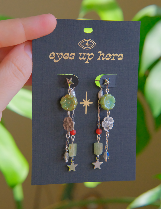 Earrings on a 'eyes up here' card held by a hand with a blurred green plant background