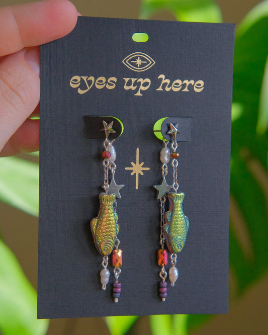 Pair of earrings on a 'eyes up here' card with a blurred green plant background