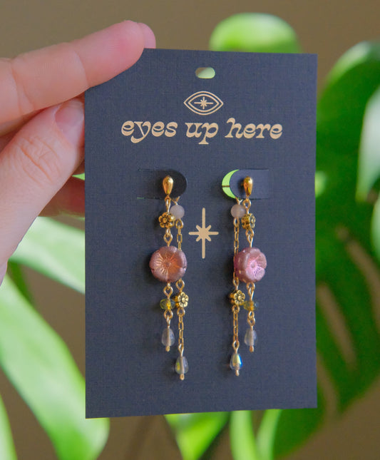 Pair of earrings on a 'eyes up here' card held by a hand with a green leafy background